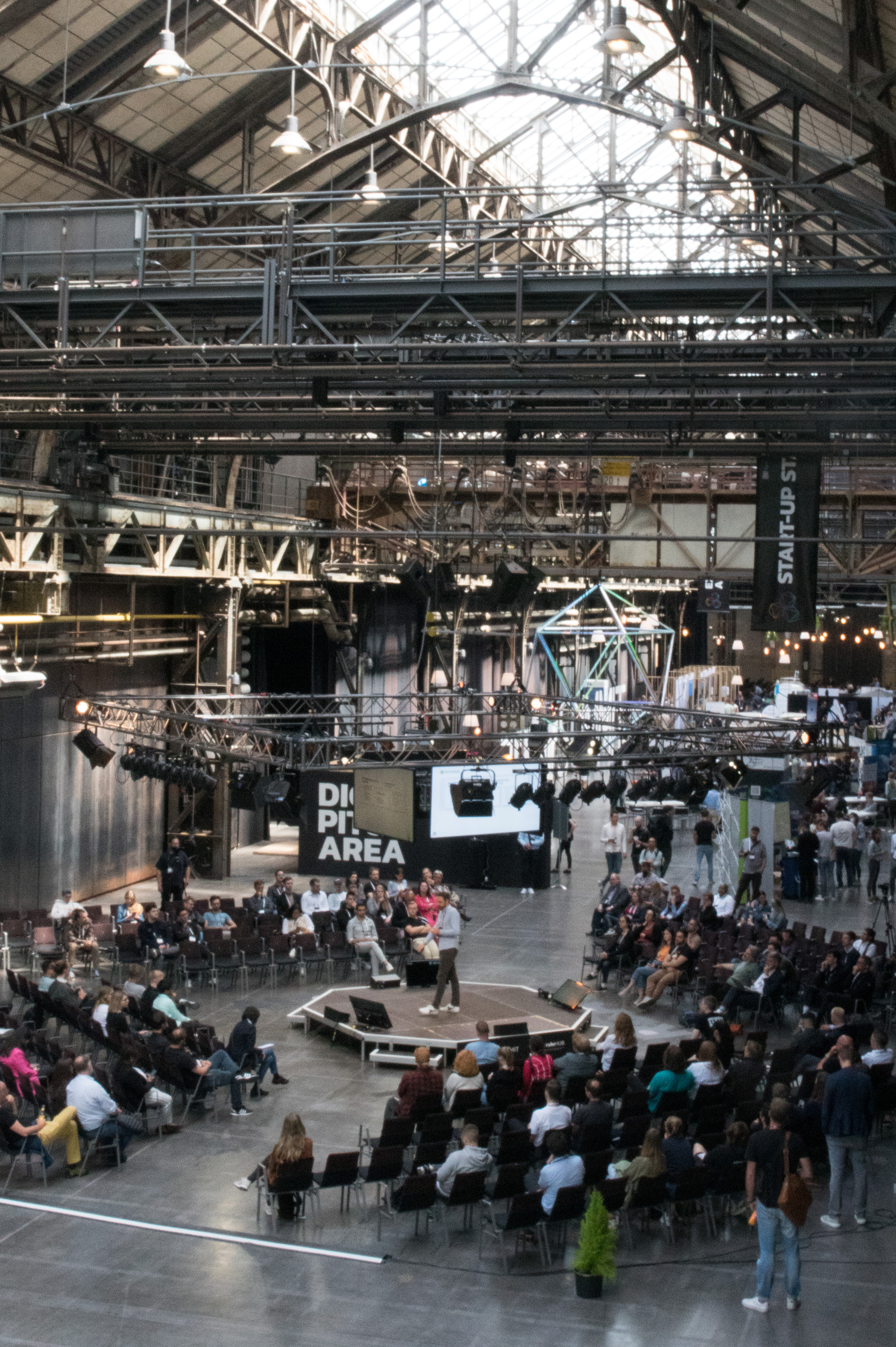 Besucher:innen des Ruhr Summit in der Halle 1 der Jahrhunderthalle Bochum sitzen um eine runde Bühne herum und lauschen einem Moderator. Die Halle ist mir Tageslicht beleuchtet.
