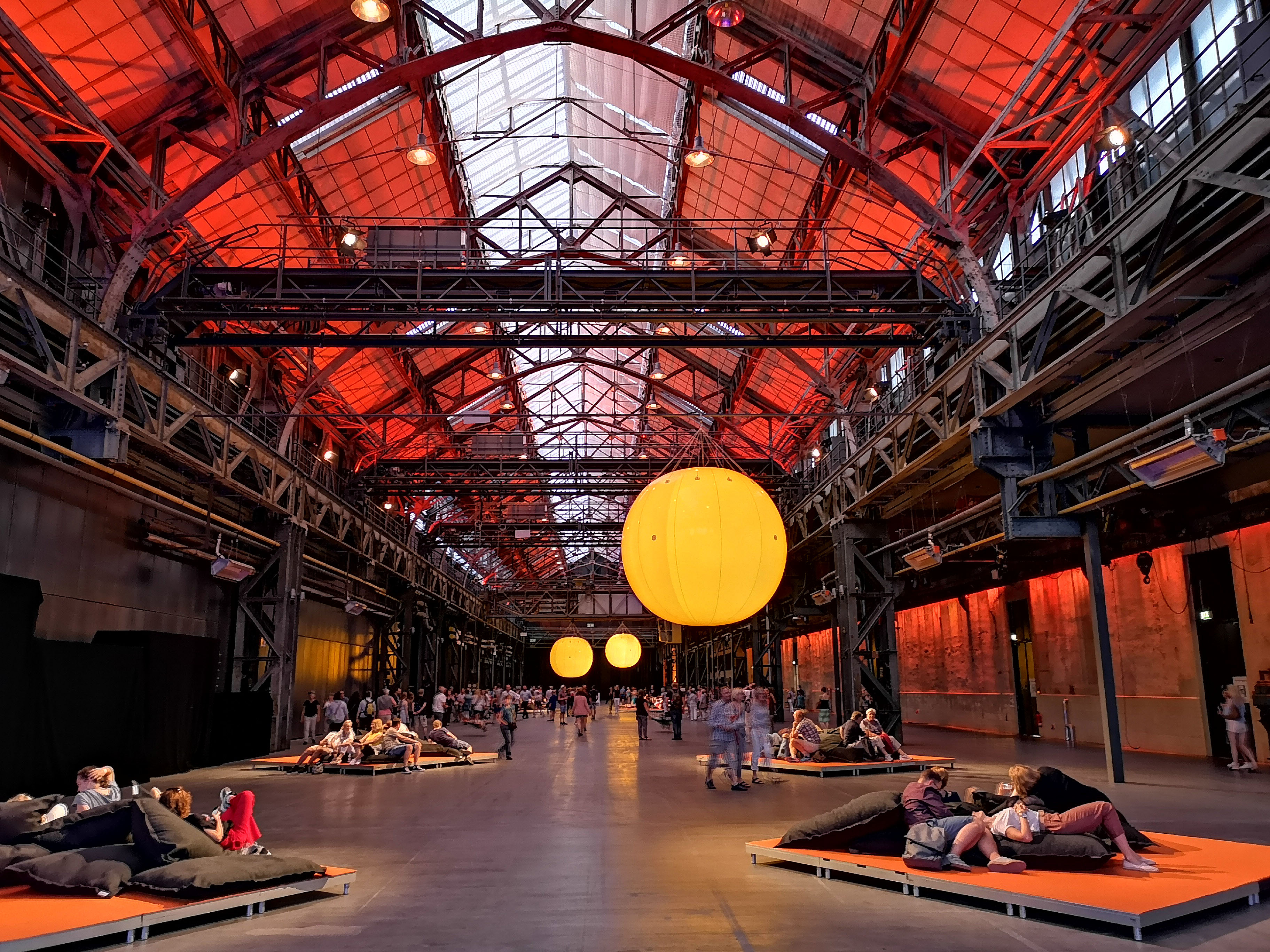 Menschen sitzen und liegen entspannt auf in der Halle verteilten Sitzmöglichkeiten und genießen die Atmosphäre der rot beleuchteten Halle 1 der Jahrhunderthalle Bochum während der ExtraSchicht. Von er Decke hänge große, gelb beleuchtete Luftballons.
